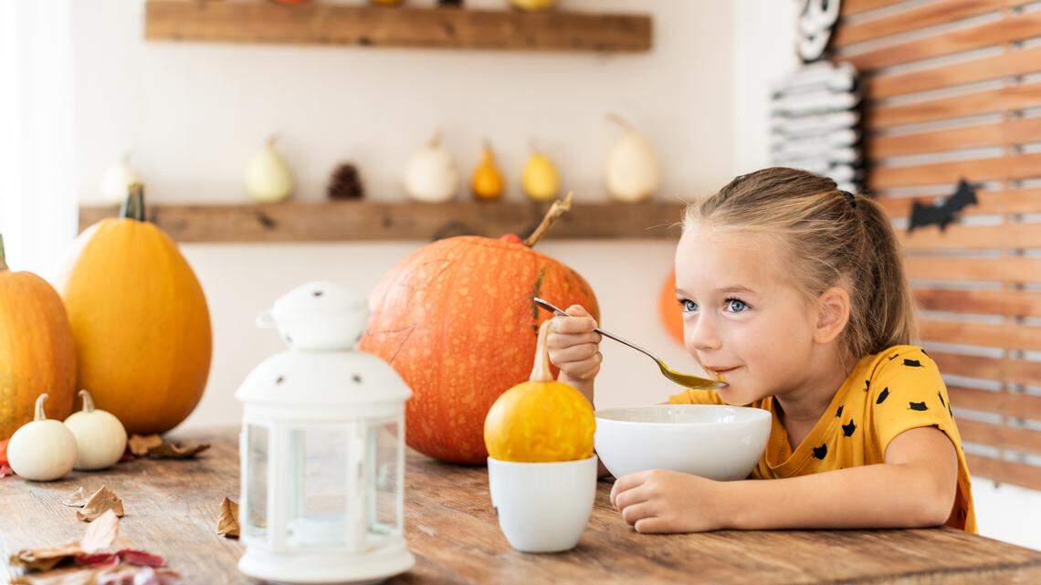 Cute little girl eating pumpkin soup in Halloween decorated dinning room. Autumn season food lifestyle background.