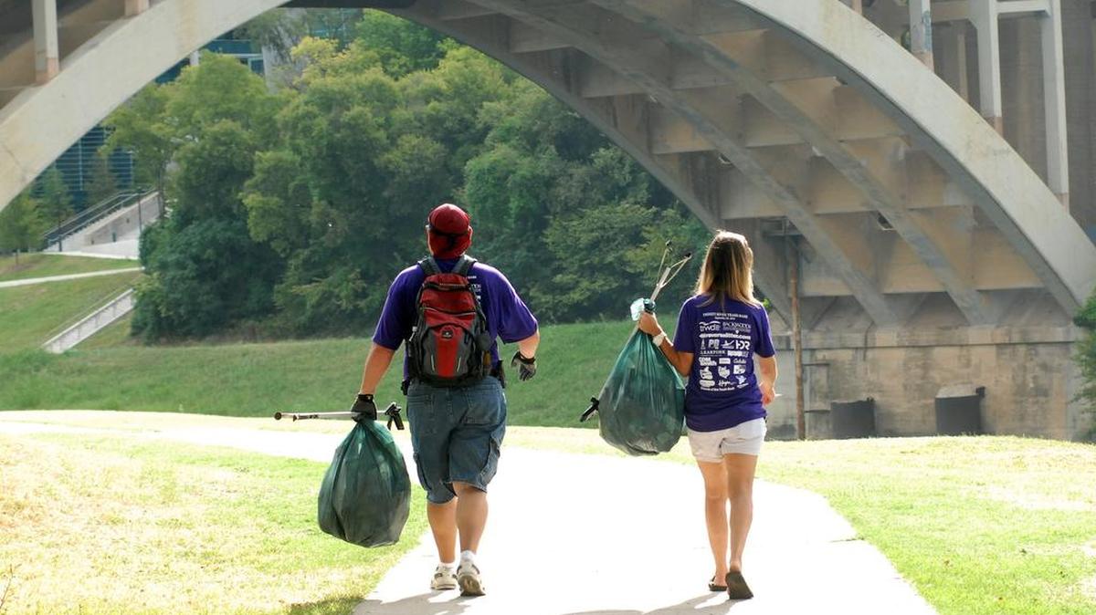 A man and woman walk on a Trinity Trail sidewalk, holding a trash bag in their left hands. Their backs are facing the camera.