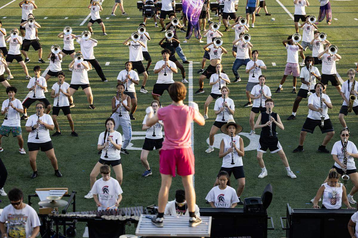 Members of the marching band stand in rows on a football field while a drum major stands in front of them on a stand.