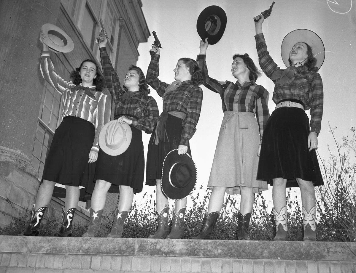 The girls are standing on a ledge, dressed in western shirts, skirts, and cowboy boots. They are waving their cowboy hats in the air as Nell, Virginia, and Sallye fire their pistols into the air.