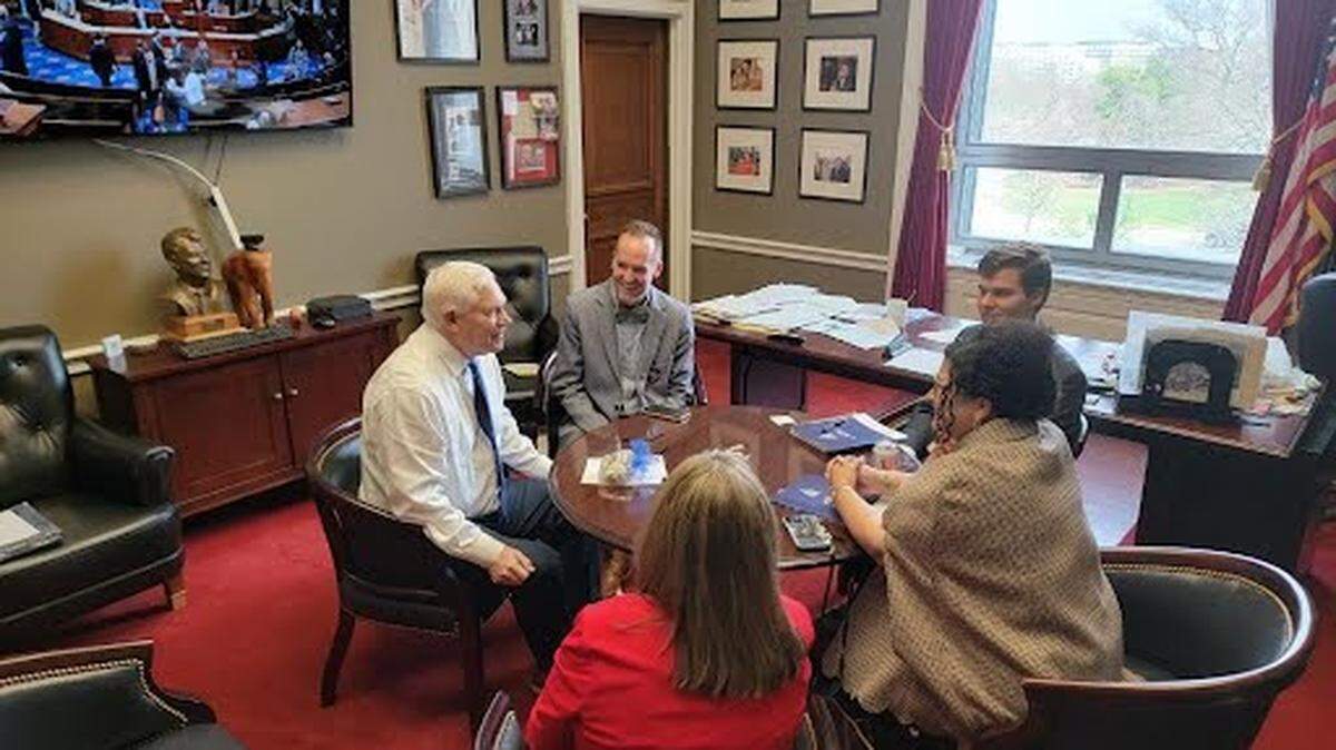 Five people sit in a circle around a table in a red-carpeted Congressional office.