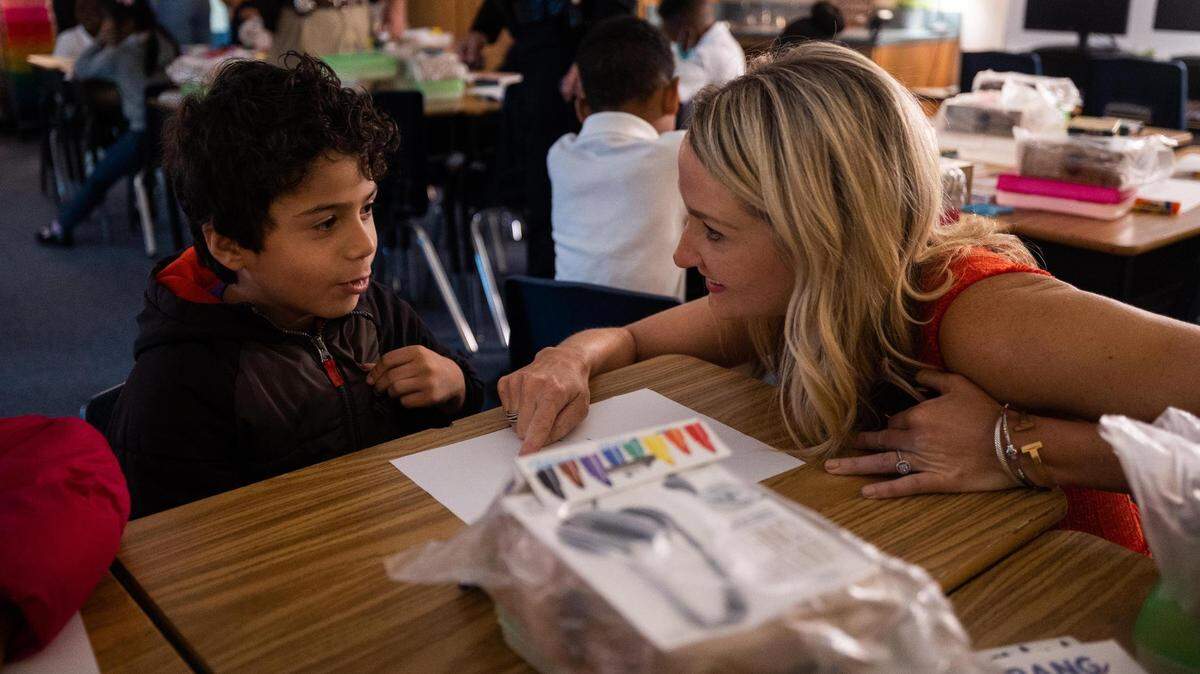 A woman crouches down to talk to a child in a crowded elementary school.