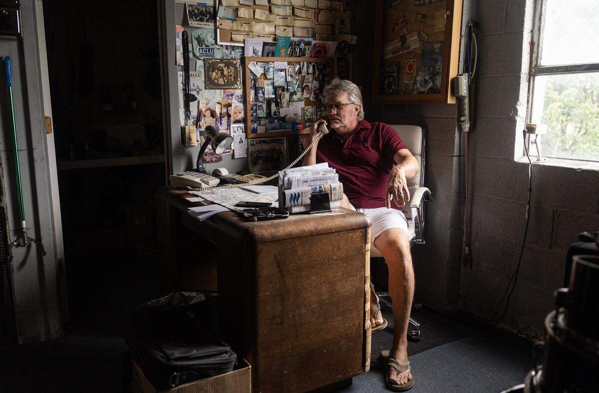 An older man sits at his desk lit by harsh window light. The back wall is covered with old paper and bulletins.