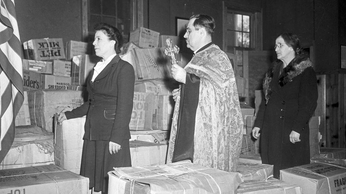 Sharing the bounty of American life, the Rev. Anastasios Vlamides, pastor of Saint Demetrios Greek Orthodox Church, blesses cartons of war relief aid bound for Greece, accompanied by Rosa Dear, left, and Nora Heras.