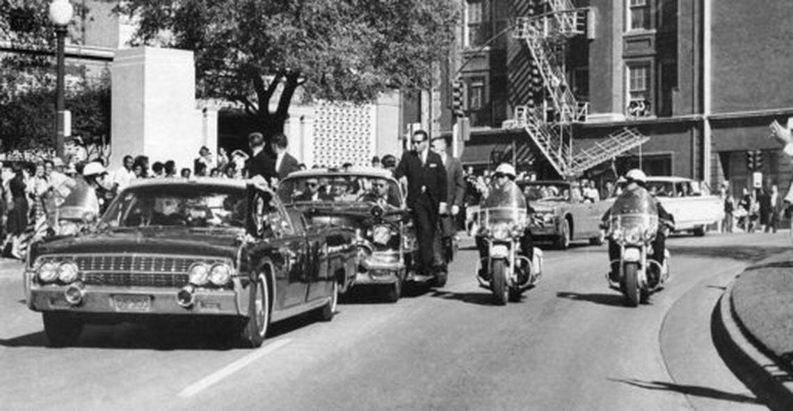 In this Nov. 22, 1963, file photo, seen through the foreground convertible’s windshield, President John F. Kennedy’s hand reaches toward his head within seconds of being fatally shot as first lady Jacqueline Kennedy holds his forearm as the motorcade proceeds along Elm Street past the Texas School Book Depository in Dallas. (AP Photo/James W. “Ike” Altgens, File)