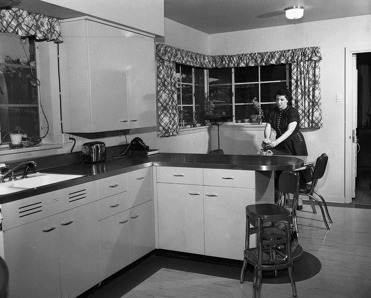 April 21, 1951: The kitchen at the home of Mr. and Mrs. Elmo Webb at 3866 South Hills Circle. Mrs. Webb is shown in front of the corner window, which overlooks the big front yard. Metal chairs and table furnish the breakfast nook beyond the service bar. The Webb home, designed as a rambler by Charles E. Armstrong, architect, was furnished for comfortable living.