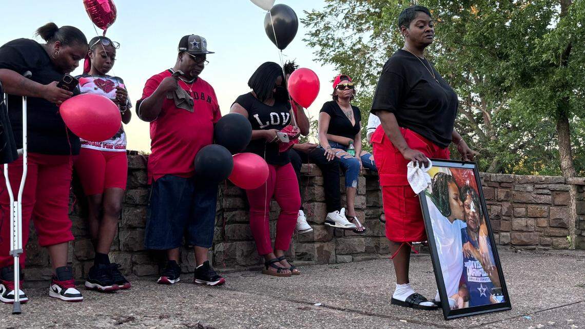 A woman in a black shirt and red pants holds a large photo of herself with a younger woman. She has a downcast look on her face. Behind her, others dressed in red and black sit on a low stone wall holding red, black and white ballons.
