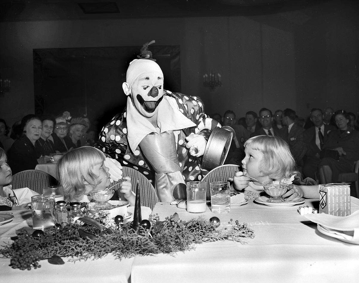 A clown bends down to a banquet table where two little girls stare at his smiling face with mystified expressions.