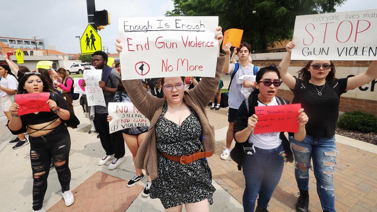 Teenagers stand on a sidewalk holding signs reading "Enough is enough - end gun violence" and "Stop gun violence."