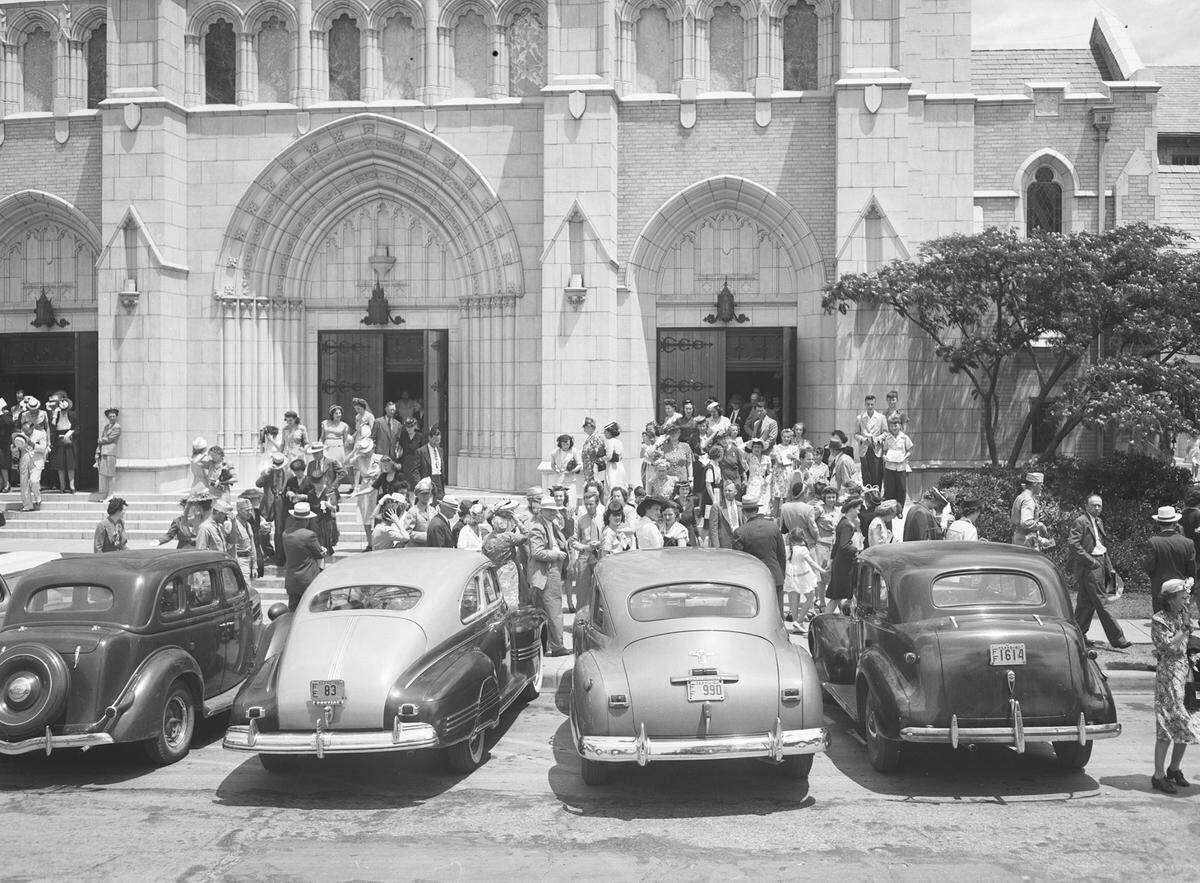 First Methodist Church is dedicated on June 10, 1945. Part of the large crowd that attended the dedication are filing out of the packed auditorium. They are standing out front of the church, mingling. There are several automobiles parked at the curb.