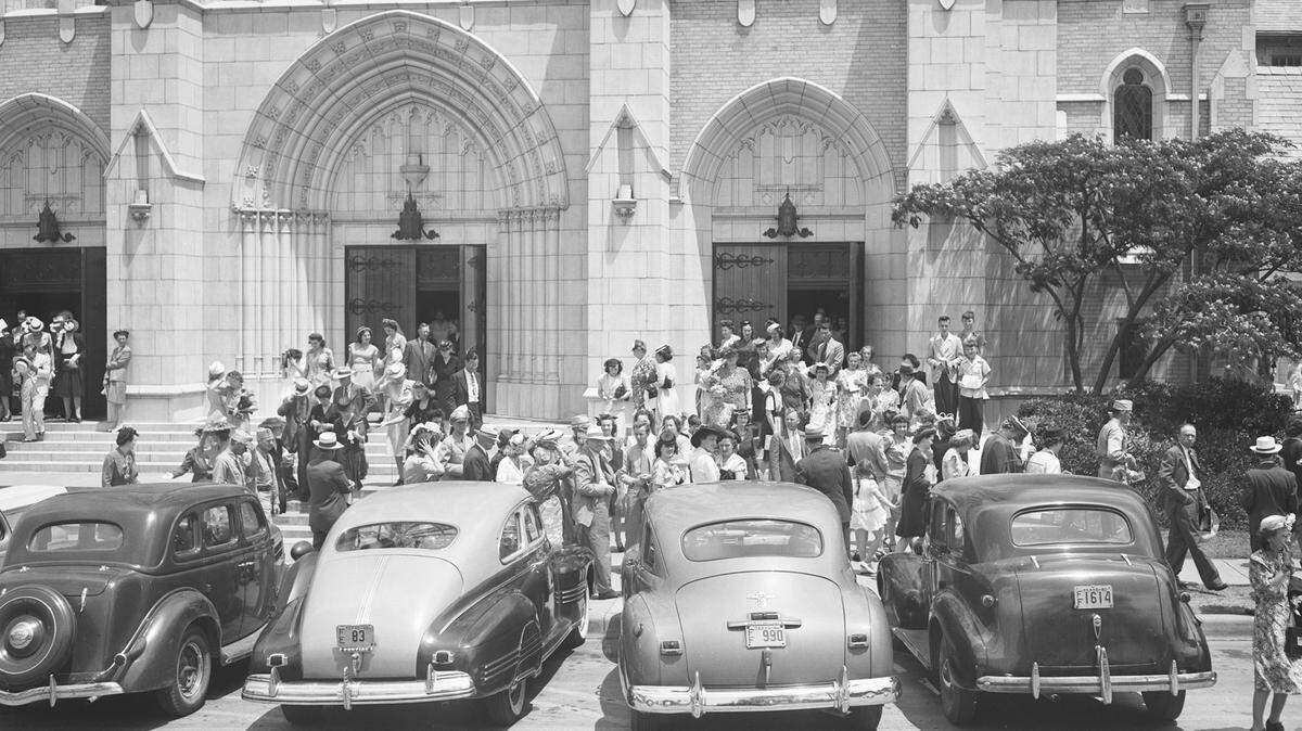 First Methodist Church is dedicated on June 10, 1945. Part of the large crowd that attended the dedication are filing out of the packed auditorium. They are standing out front of the church, mingling. There are several automobiles parked at the curb.