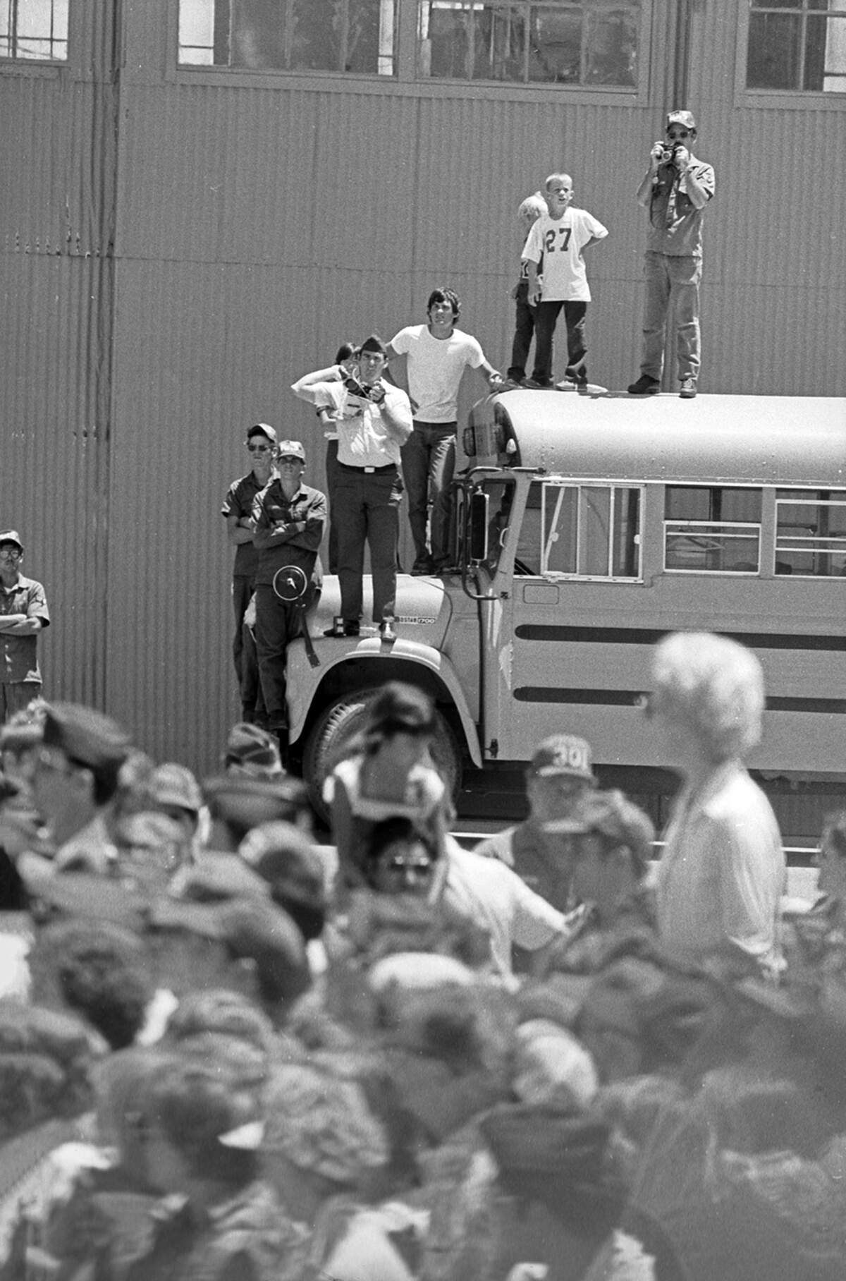 Men, including some in military fatigues, stand on the hood and roof of a parked school bus to see over the crowd. Some are holding cameras.