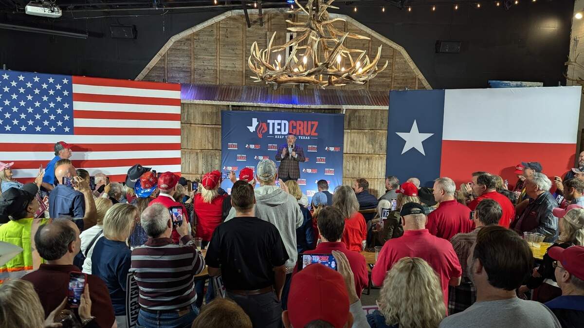 A man standing on stage between an American flag on the left and a Texas flag on the right talking to a group of around 100 people