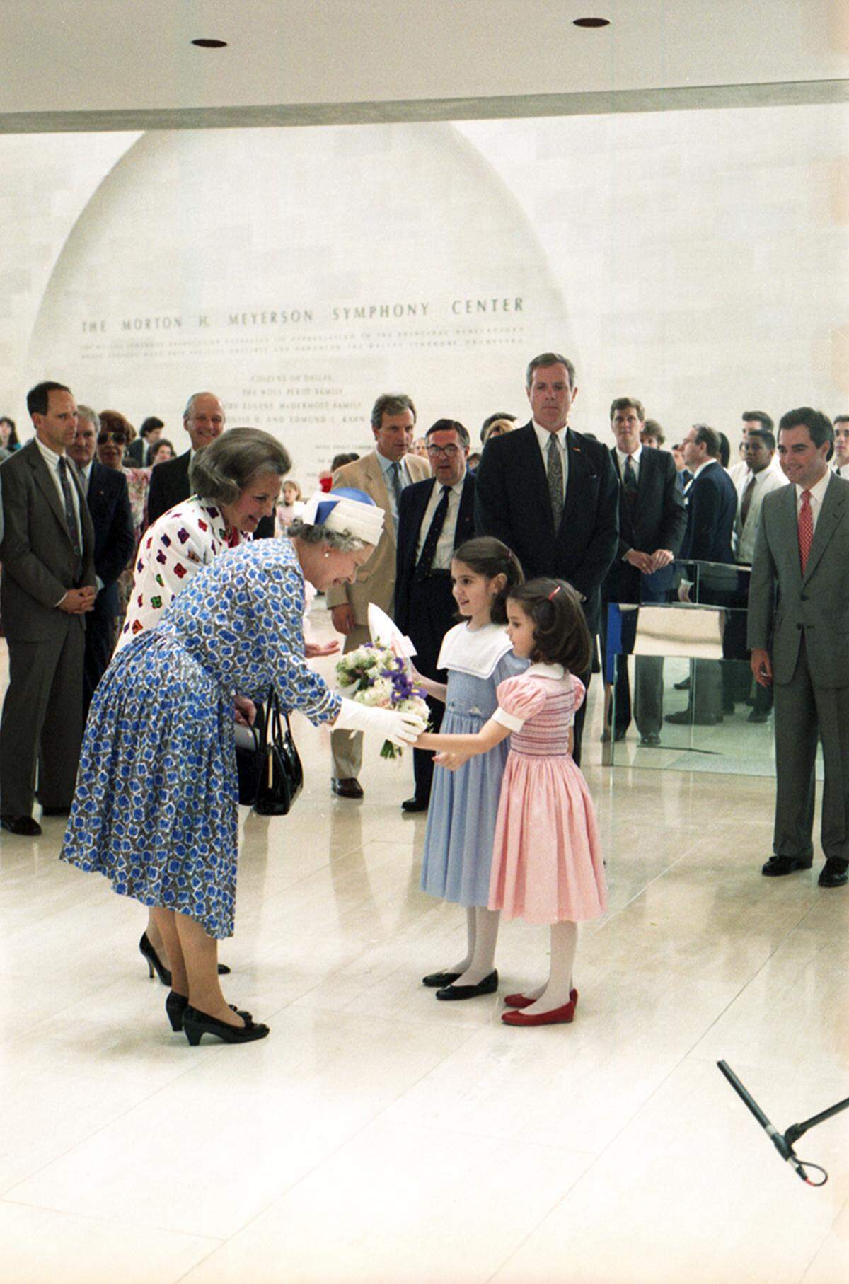 Queen Elizabeth bends down to accept flowers and a gift from two young girls at the Morton H. Meyerson Symphony Center in Dallas on May 21, 1991. Dallas Mayor Annette Strauss seen on her left. 
