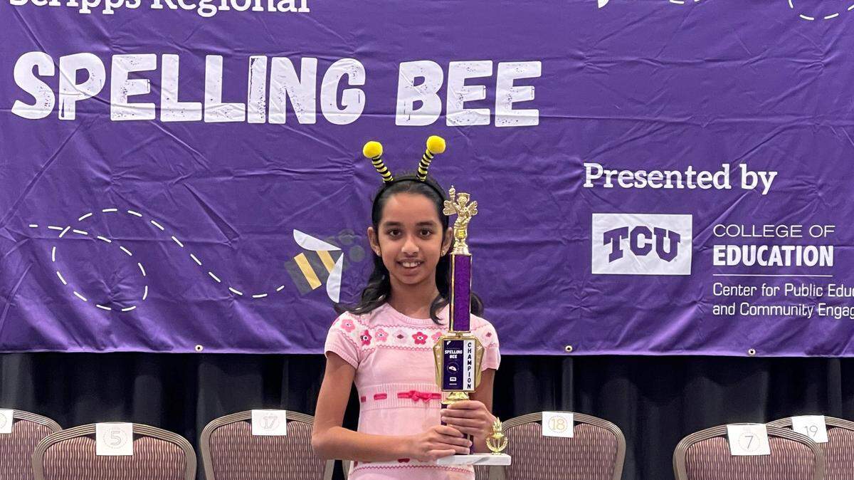 A fifth-grade girl holds a trophy in front of a Scripps Spelling Bee banner.