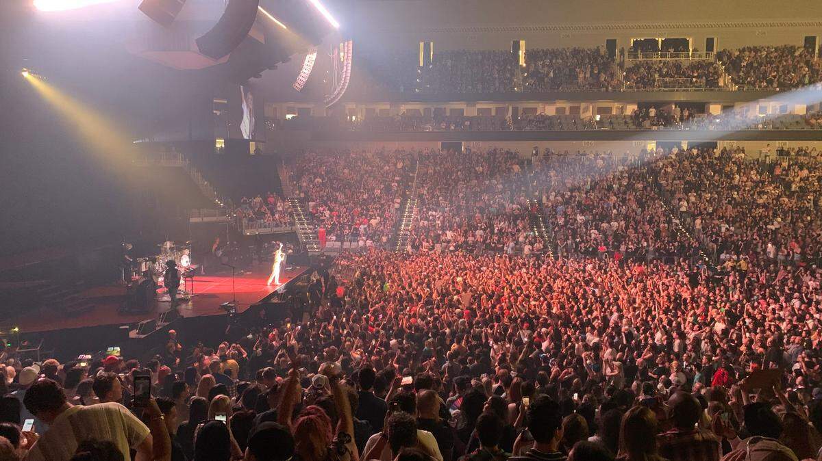 A shot of an arena concert from stage right. A packed crowd stands while a spolight shines on the lead singer, standing on the edge of the stage dressed in all white. Speakers and a light fixture hang from the ceiling.