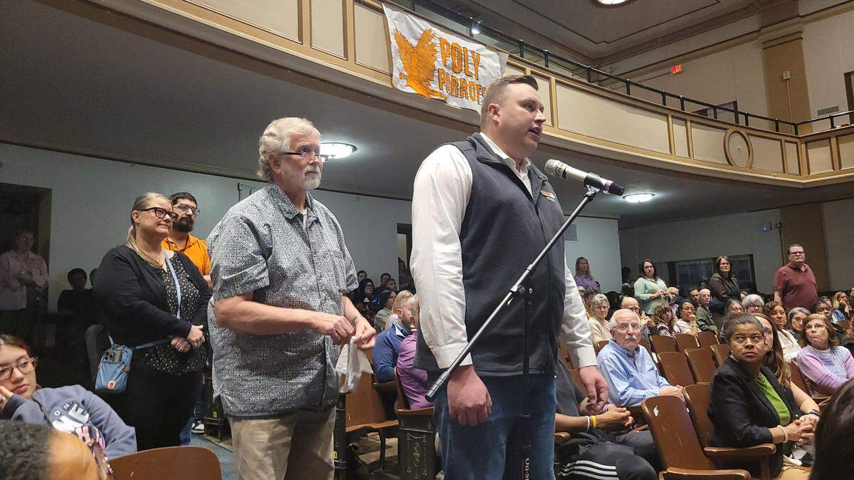 A man stands at a microphone and speaks while others line up behind him. Above them is a banner that reads “Poly Parrots,” the school’s mascot.