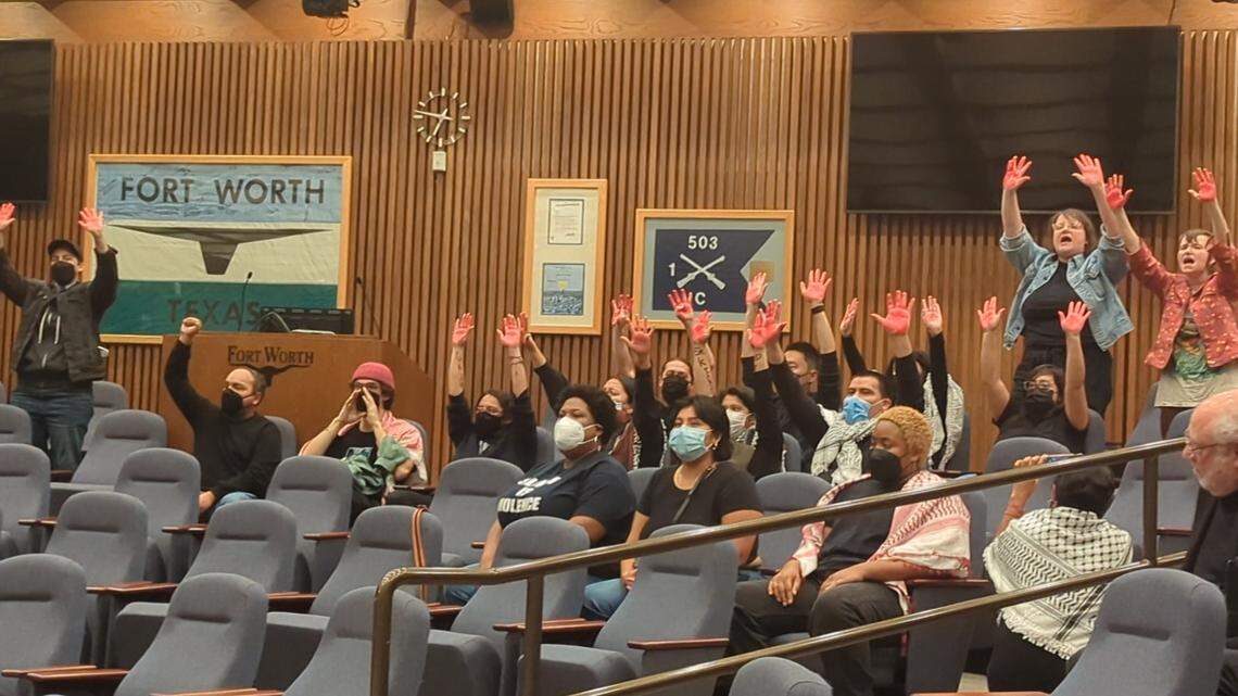 A group of people raising their hands stained with red paint while chanting in a city council chamber. 
