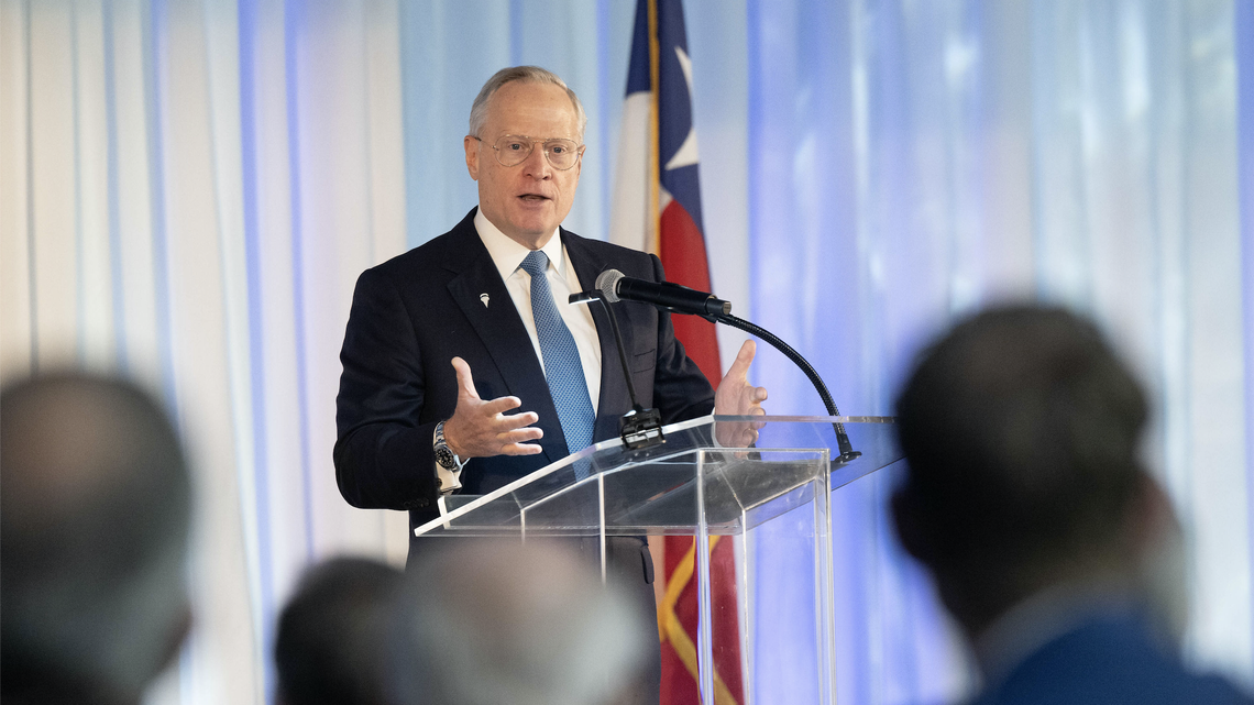 Ross Perot Jr. stands at a glass podium with his arms out to emphasize his remarks. He wears a business suit and stands in front of an American flag. 