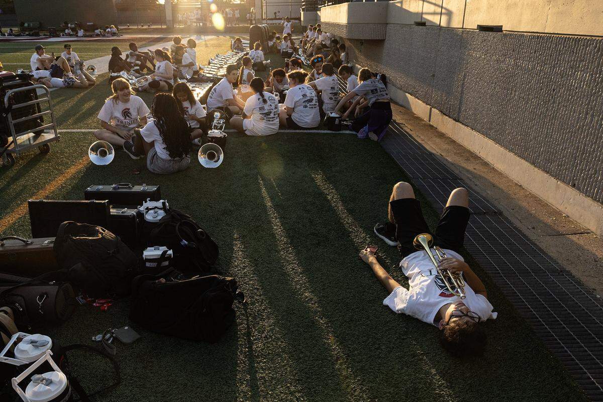 Trumpet player Addan Barber, 17, lays on the ground with his trumpet resting on his chest while his bandmates sit in a circle farther away resting.