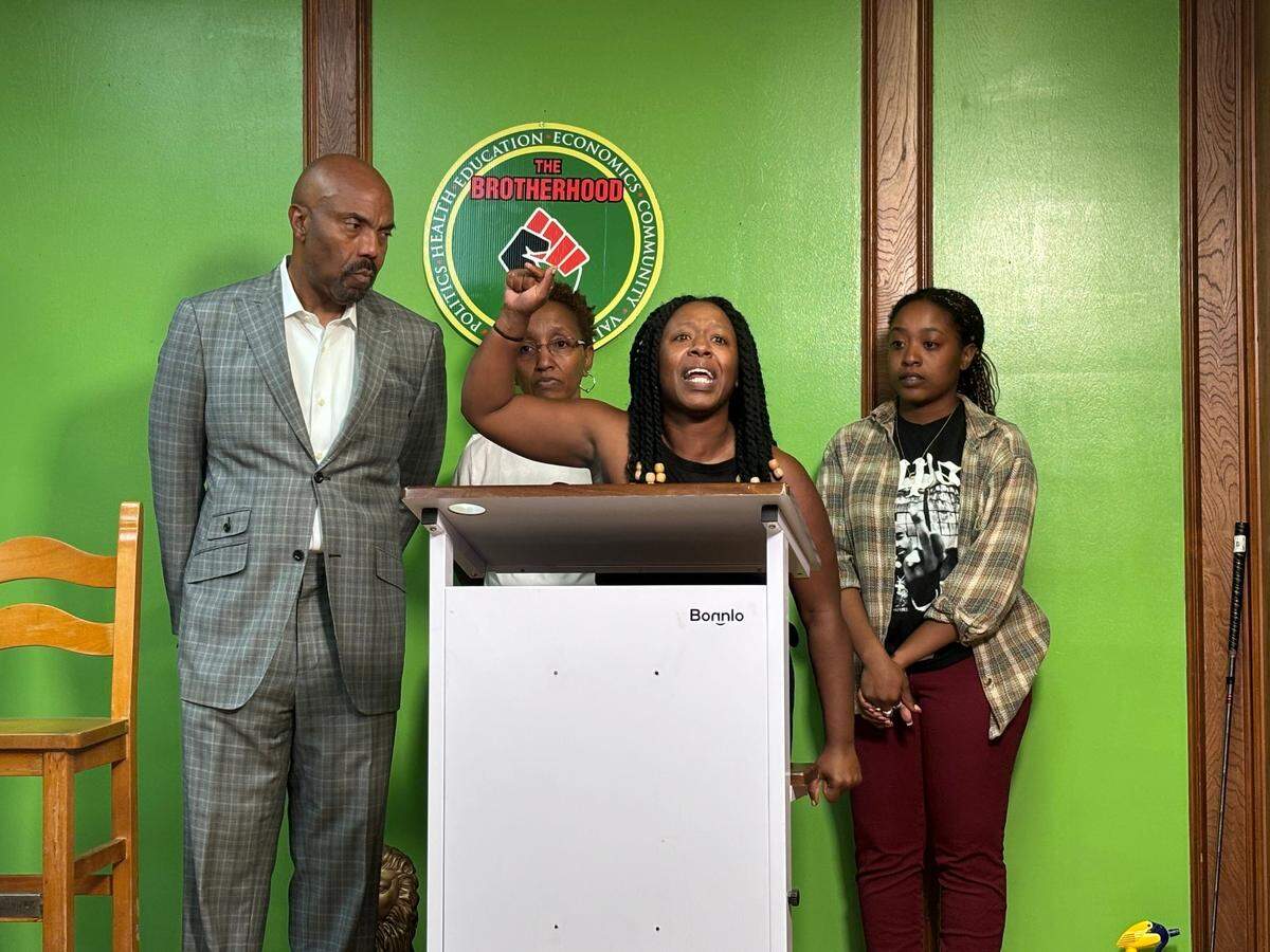 A woman with a pained look on her face raises her fist as she speaks behind a lectern. Two other women and a man in a suit stand behind her. They stand before a lime green wall. 