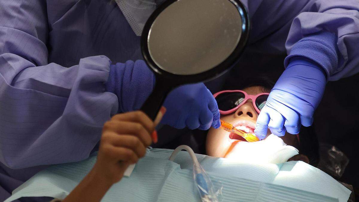 A little girl lays back with a bib covering her body and heart-shaped eyeglasses protecting her from bright light as a dental student in scrubs cleans the girl's teeth.