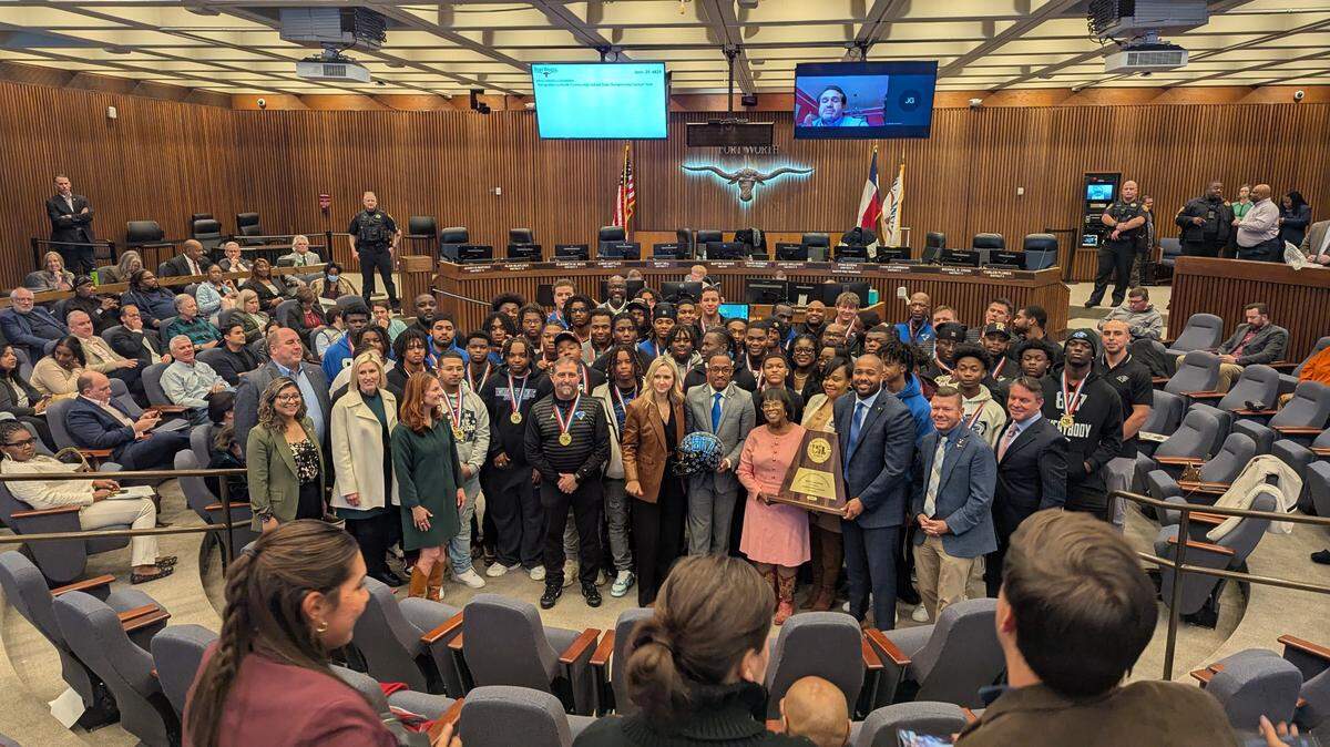 A group photo of a high school football team in a city council chamber surrounding a championship trophy