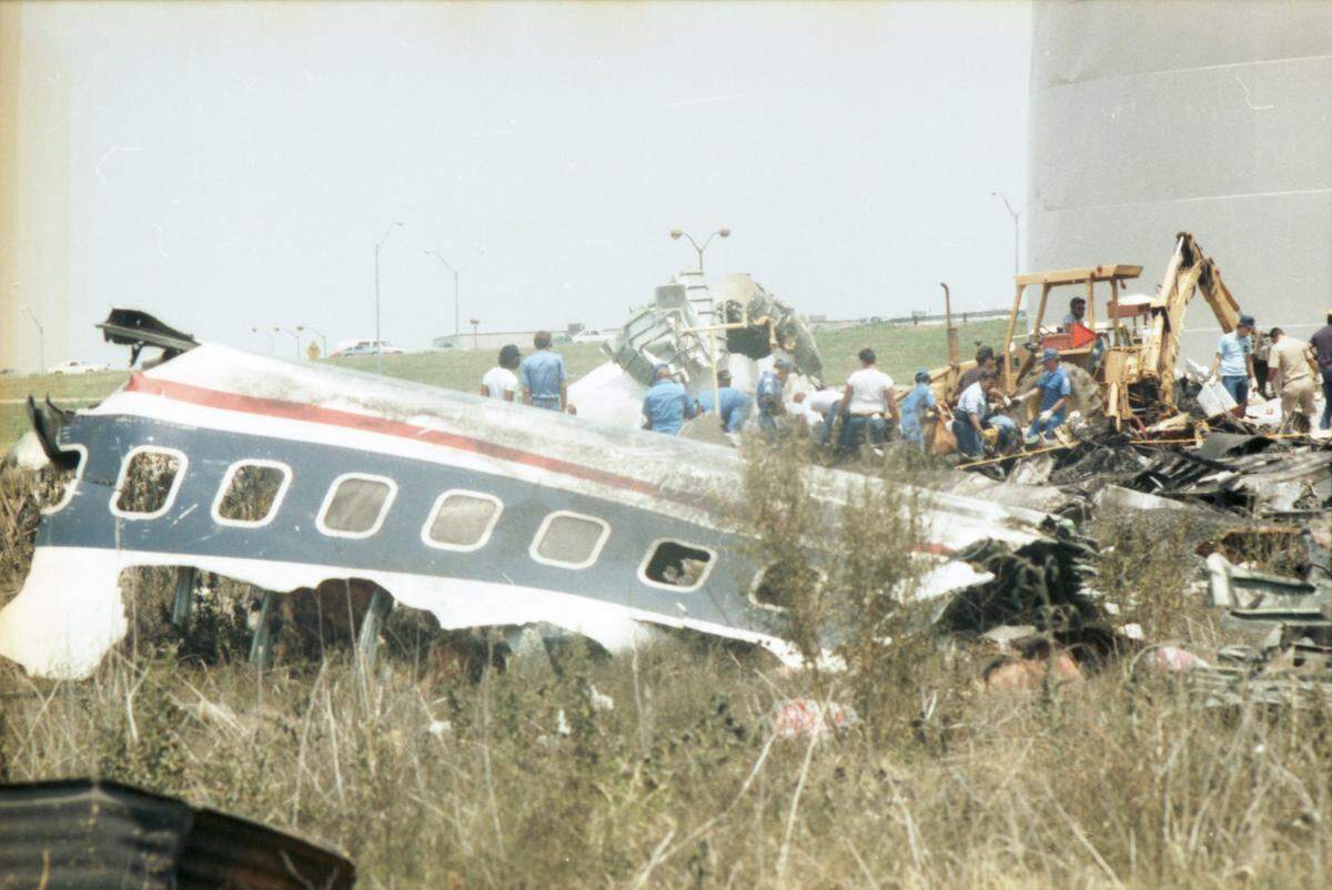 A large section of plane wreckage in a field with emergency workers in the background.