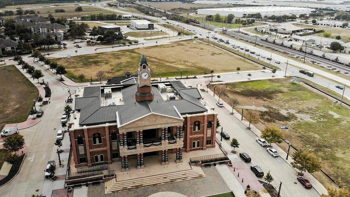 A aerial photograph showing a large two-story building in the foreground with vacant grass plots behind and to the right of it.