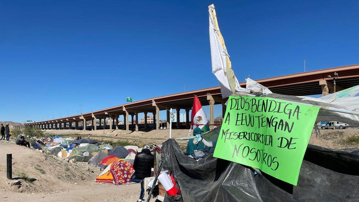 A green poster board sign hangs outside a tent made of black tarp. A white flag flies overhead. Tents stretch out to the left of the makeshift shelter and a highway across the river beyond looms overhead. 