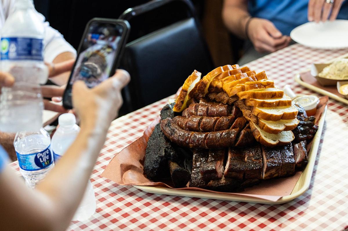 A customer takes a photo of a large tray of barbecue stacked with Lao sausage, pork ribs, beef ribs and freshly baked sliced bread at Goldee’s BBQ south of Fort Worth on Friday, June 28, 2024.