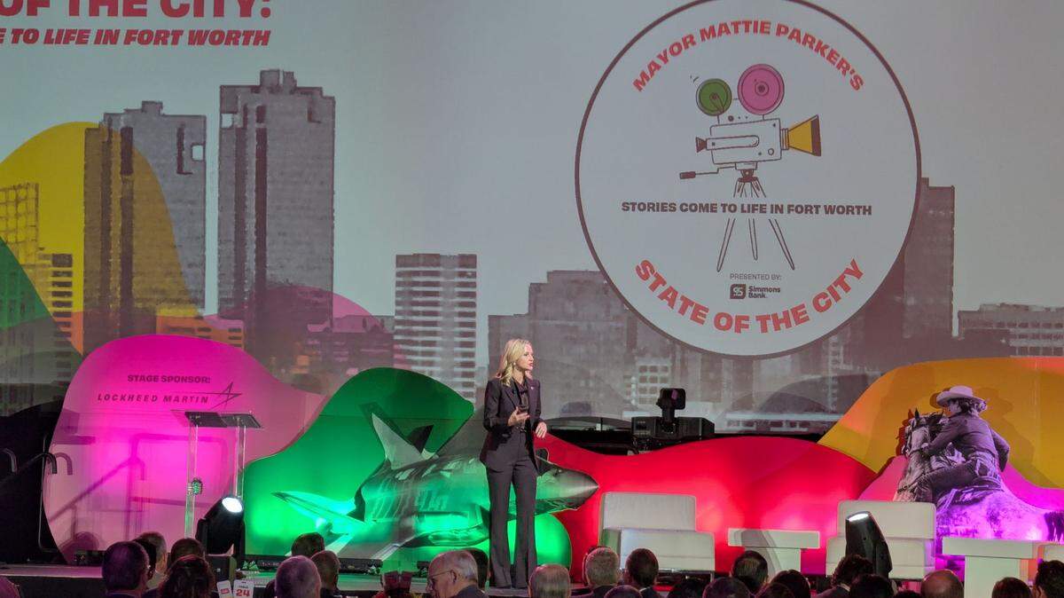 A blond lady stands up on a stage talking to people about how great Fort Worth is