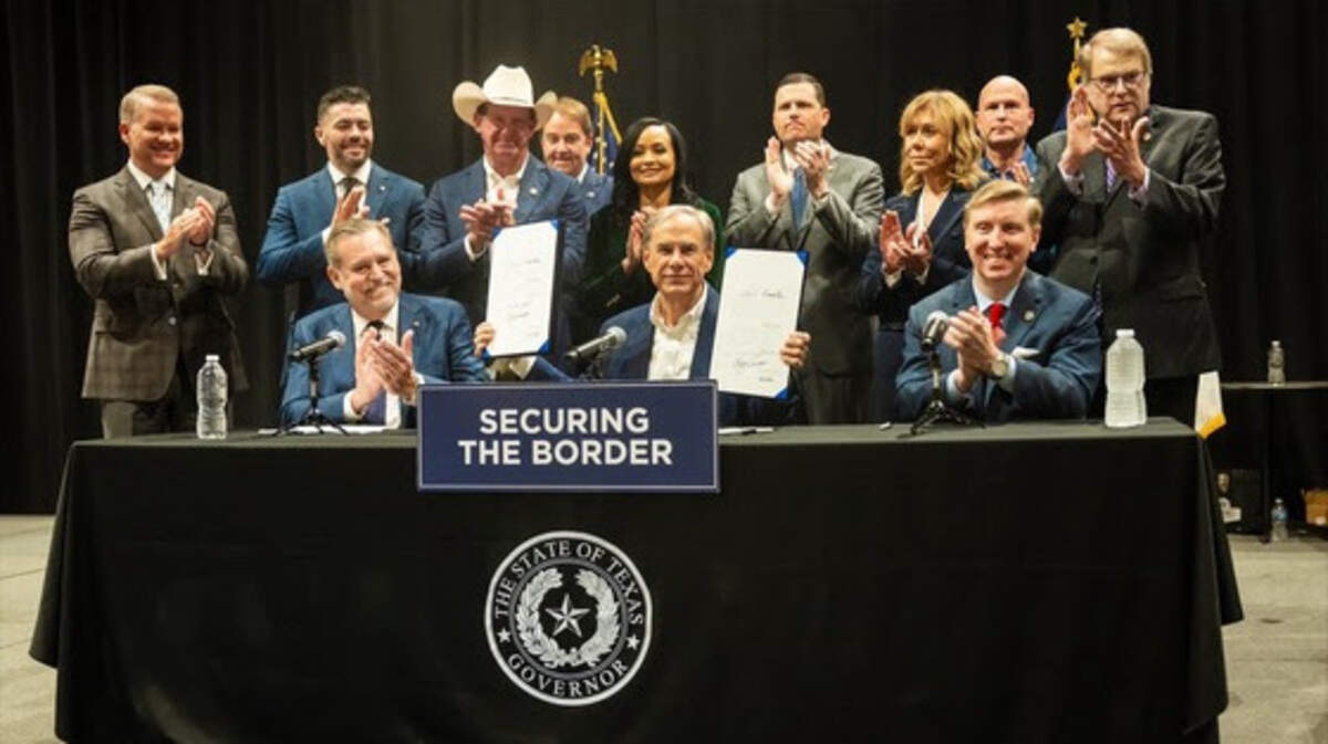 A man sitting behind a table holding up two signed bills while surrounded by applauding state lawmakers. 