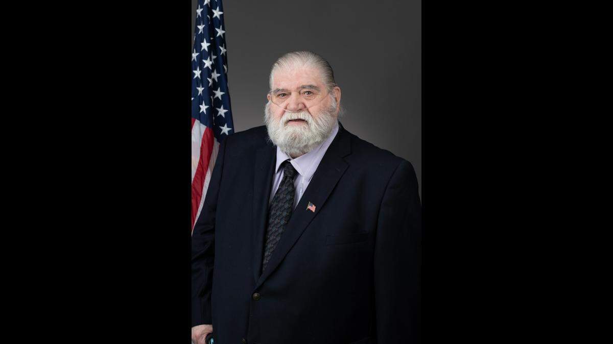 An older white man man in a black suit with an oxygen tube under his nose standing in front of a grey background with an American flag to his left.
