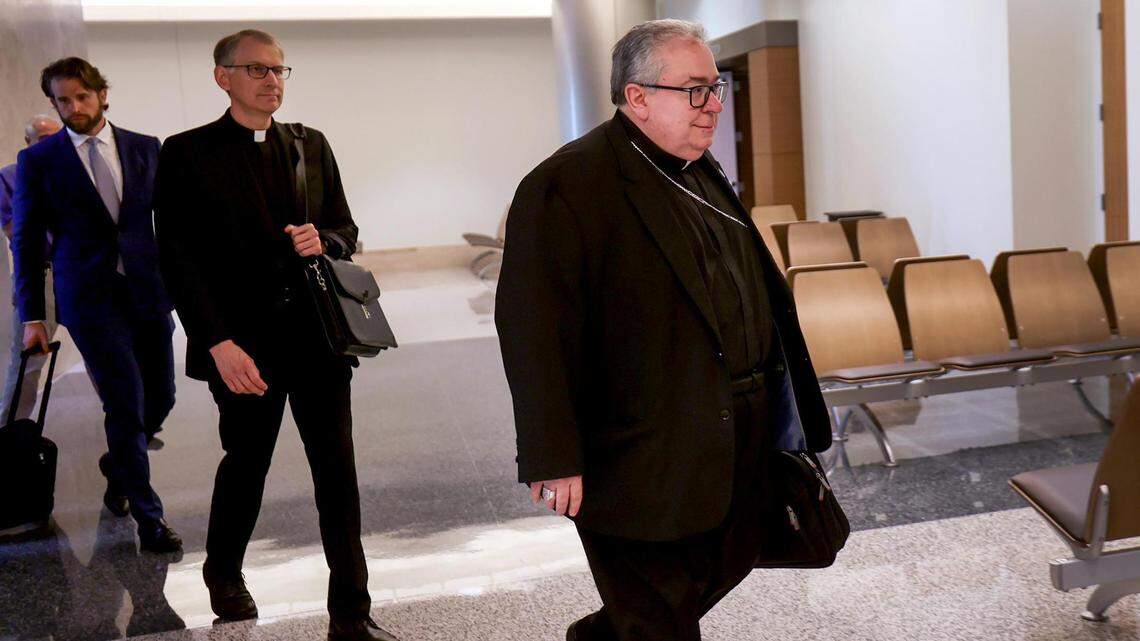 The bishop, dressed in black with a clerical collar and chain, walks in the hallway to a courtroom followed by a priest and man in a suit with rolling briefcase.