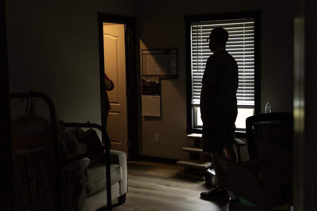 Kevin Kirkland stands in his bedroom silhouetted by a window with venetian blinds. The bedroom is dark, but to the left a light glows through the doorway.