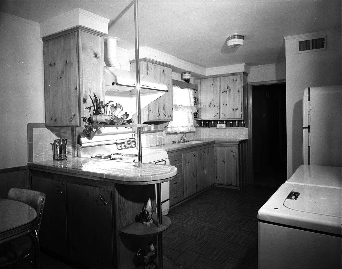 March 27, 1953: Assembly line methods are practiced in this compact kitchen of the R.J. Carlson home, 4820 Lowell Lane. There are two steps separating the refrigerator (extreme right) and the working area around stove. The drain is tiled. Cabinets are knotty pine in natural finish. It is a kitchen with wash and dryer machines in the kitchen.