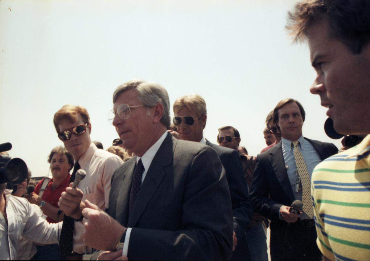 A man surrounded by people in harsh light, seeming to be talking to reporters. 