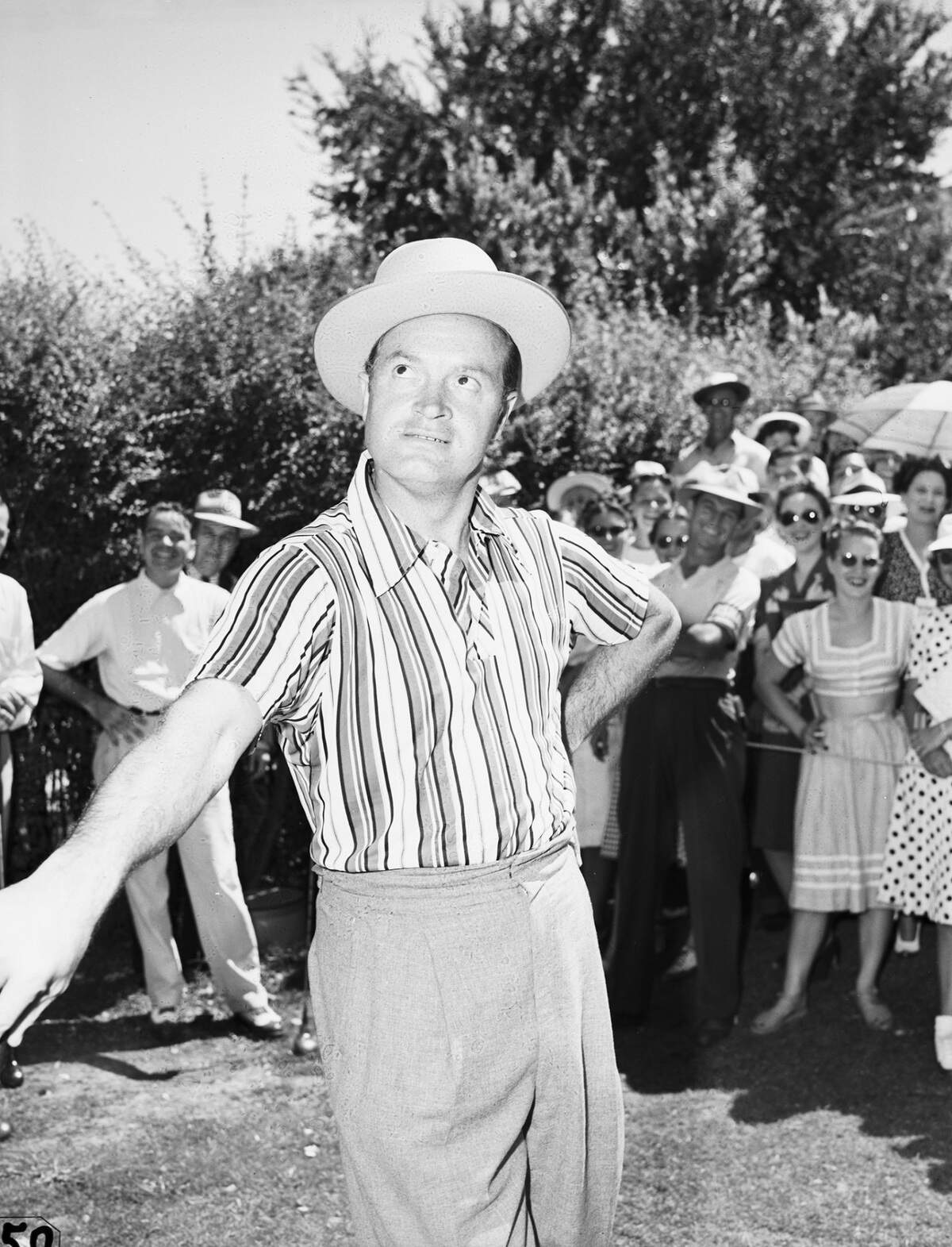 He is wearing a hat, a striped short sleeve shirt and trousers as he leans on his golf club. A crowd of people are following the famous actor as he makes his way around the course.