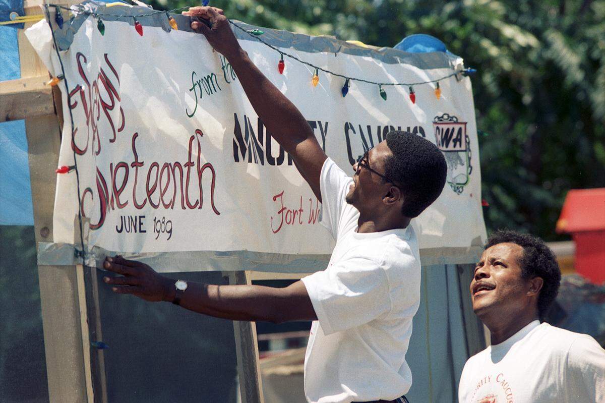 Clarence Walker, left, tapes up a sign at a Juneteenth celebration in 1989 that drew hundreds in Fort Worth. Looking on is Glen Holt. The sign reads “Happy Juneteenth from the Minority Caucus, Fort Worth, Texas, June 1989” and to the right there is a CWA logo for the Communications Workers of America; 1989-06-17 [FWST photographer Mark Gail]