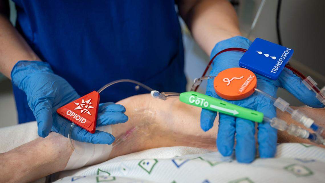 A nurse's gloved hands show four different labels on intravenous lines. One label is bright red, and says "opioid" in raised letters; another is orange, and says "antibiotic;" the third is blue, and is labeled "transfusion;" the fourth tag is green is and says "Go To," indicating an open line.