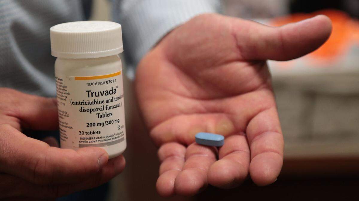 A man's hand holds a small blue pill next to a white prescription medication bottle. The bottle's lable says it contains the drug Truvada.