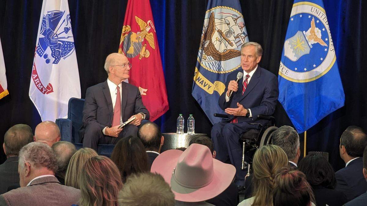 Two men sitting on a stage front of a bunch of flags from the different branches of the United States Military.