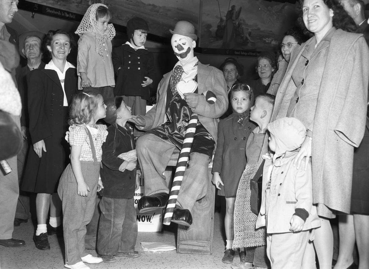 A crowd of kids and their parents gather around a large clown holding a candy cane.