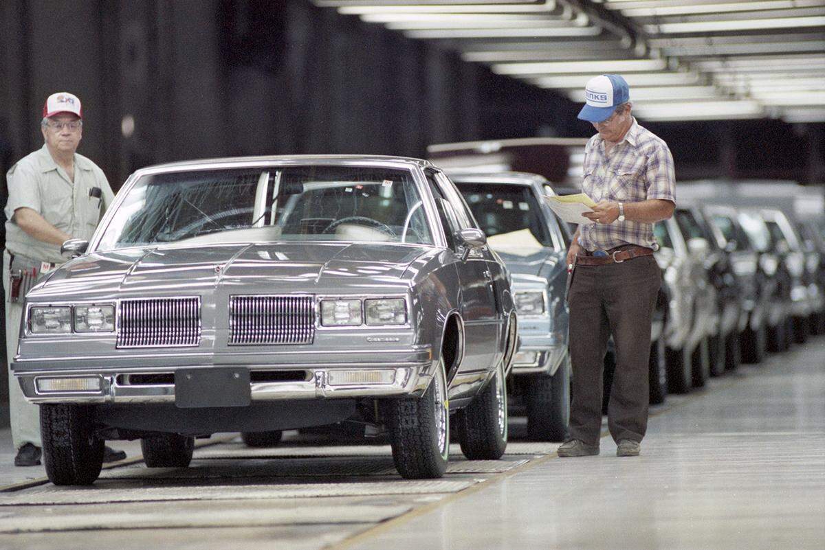 Two men wearing ball caps walk along a line of new cars on an assembly line.