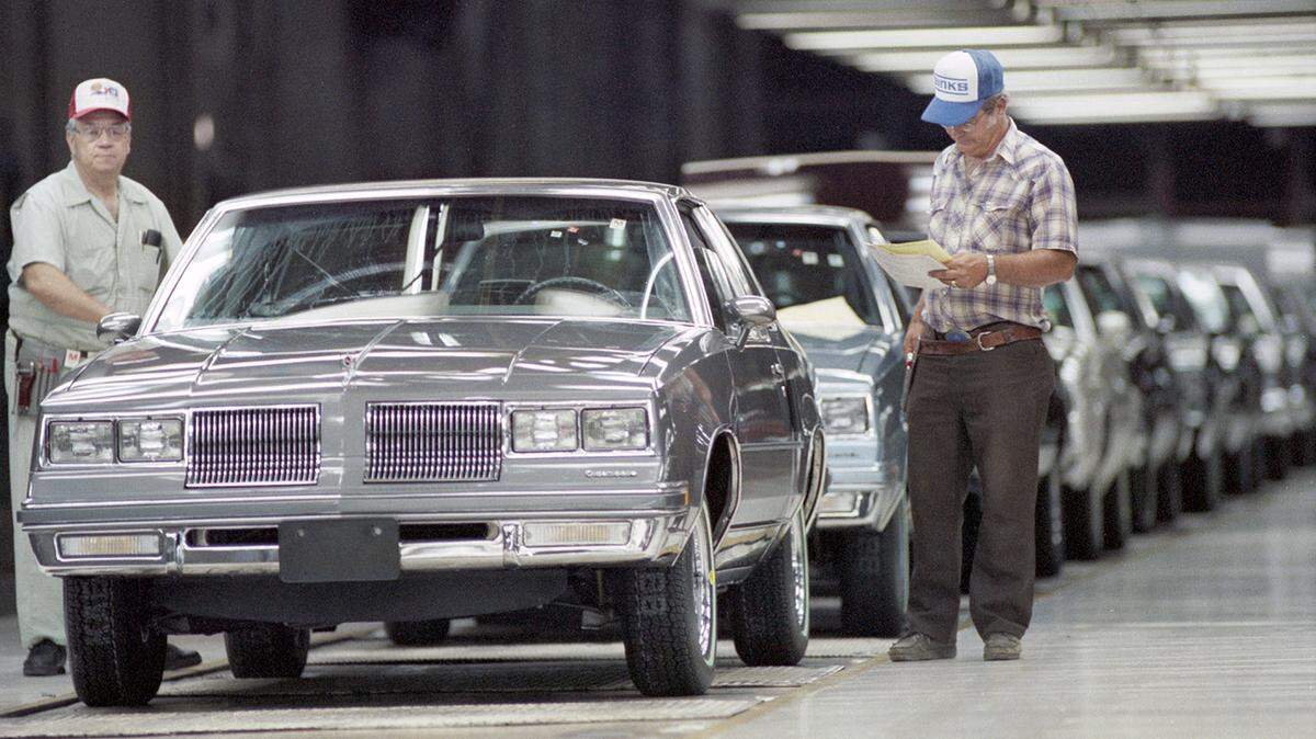 Two men wearing ball caps walk along a line of new cars on an assembly line.