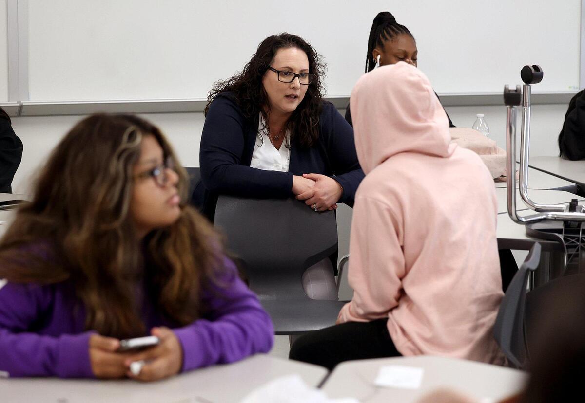 Superintendent Angelica Ramsey leans forward, while seated, to talk to a high school student in a pink hoodie in a classroom.