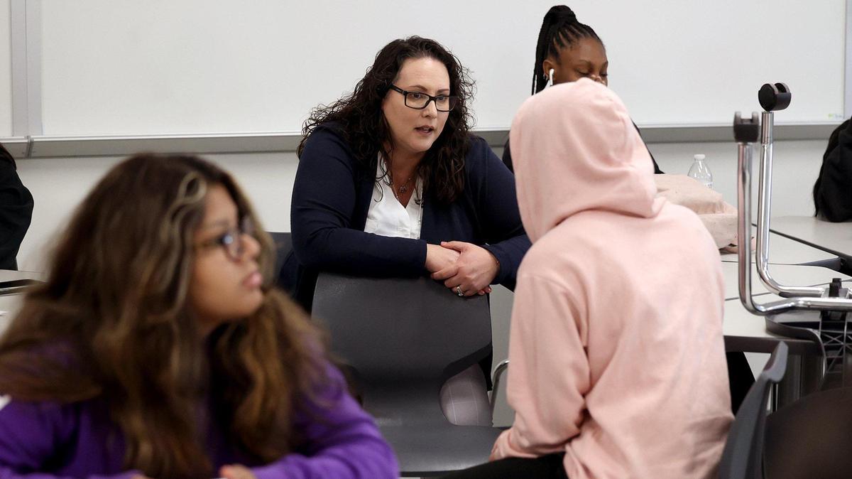 Superintendent Angelica Ramsey leans forward, while seated, to talk to a high school student in a pink hoodie in a classroom.