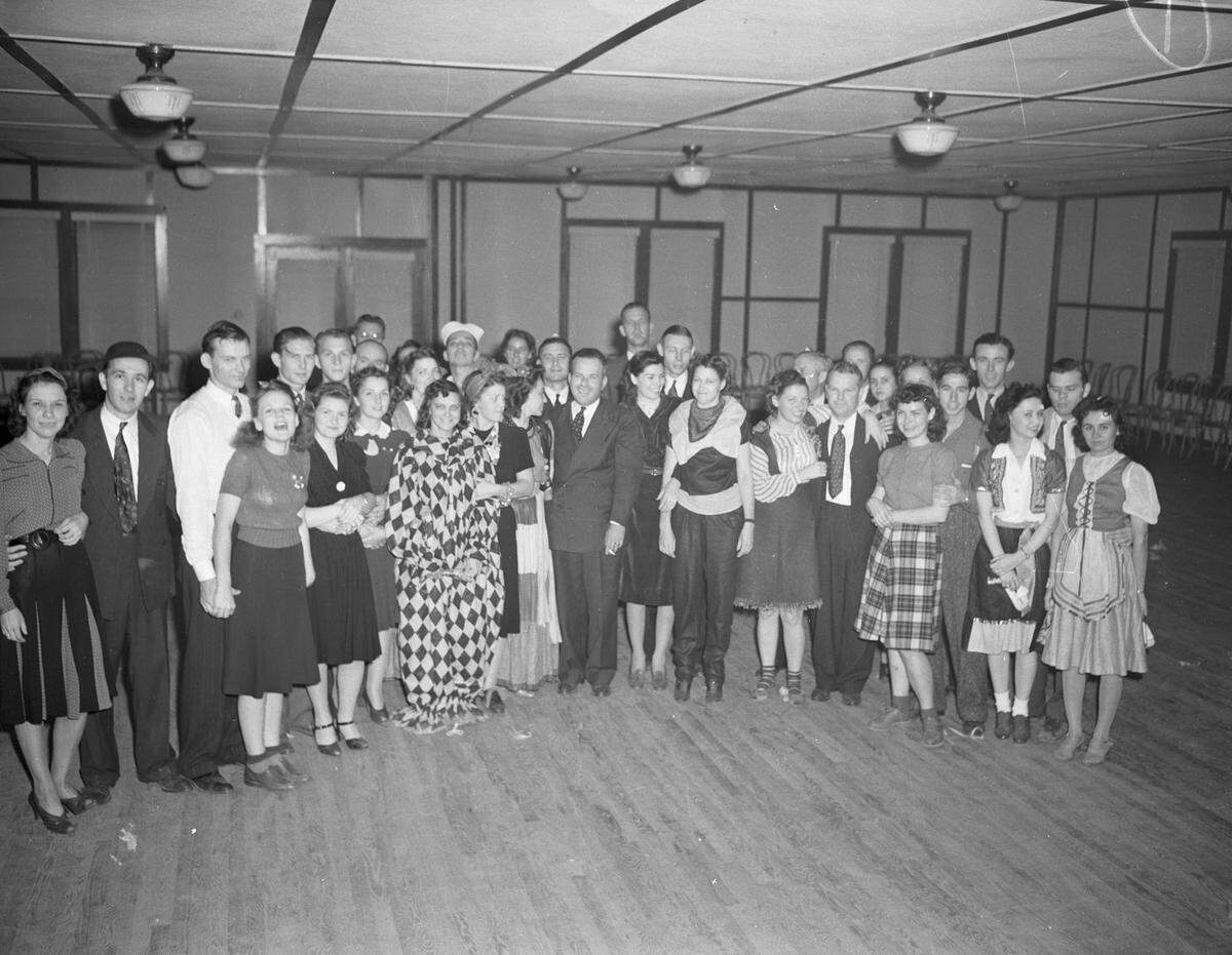 Everyone is standing grouped together in a semi-circle in an open room. Some people are in costumes. Women in the front row are wearing either dresses or blouses and skirts. Most of the men are wearing suits.