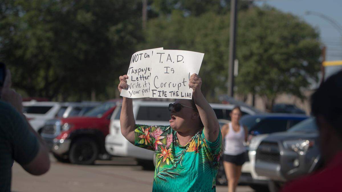 A woman holds a sign that reads "TAD is corrupt. Fire the liar"
